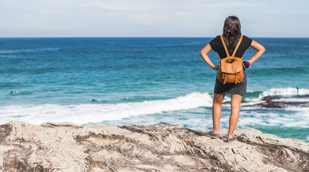 Woman with backback standing on rocks infront of blue ocean