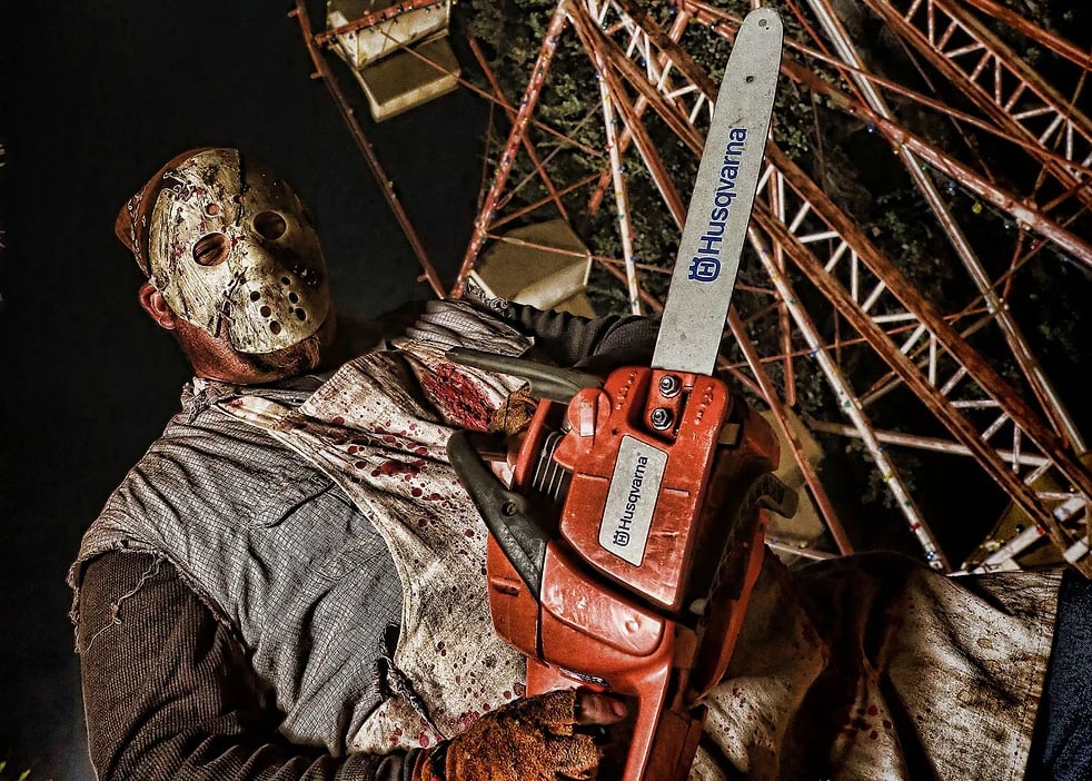 Actor in Jason Voorhees mask and chainsaw with Haunted Amusement Park in background
