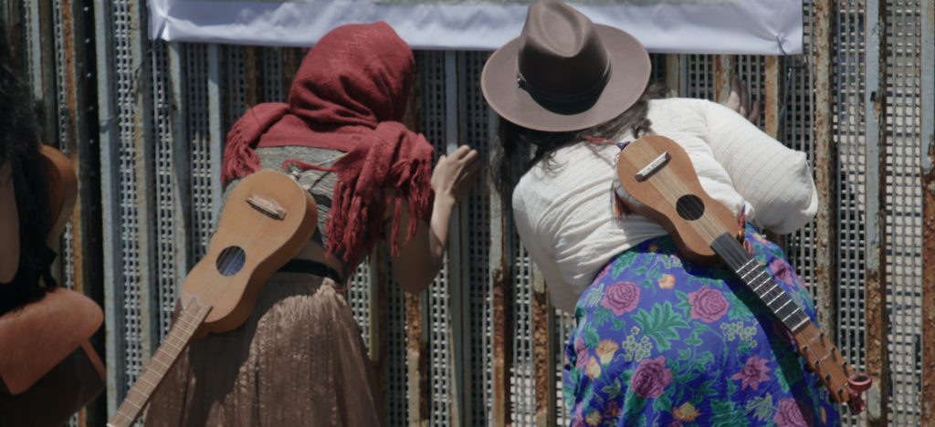 Two musicians peering through the US Mexico wall with guitars on their backs