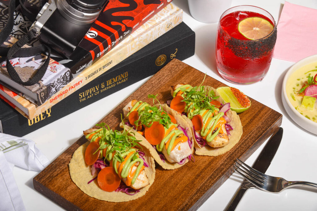 Three fish tacos on a cutting board surrounded by cocktail, camera, and Shepard Fairey books