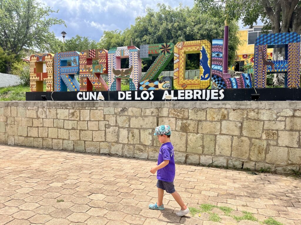 Child infront of colorful Arrazola sign with the text Cuna De Los Alberijes underneath in Oaxaca Mexico