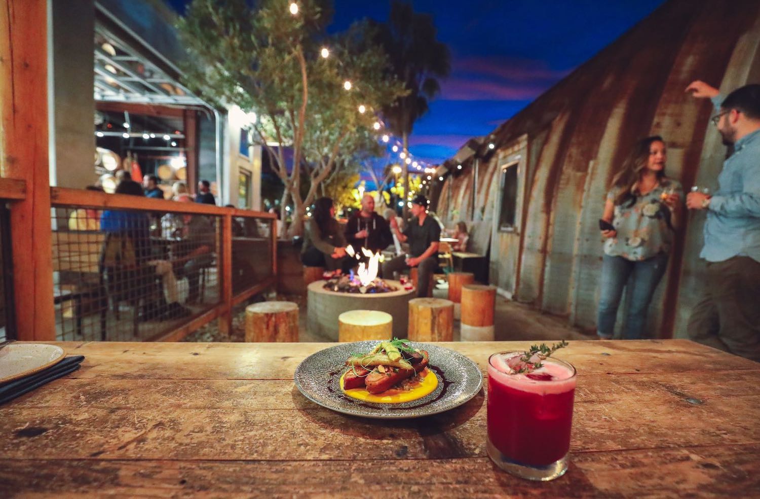 Large groups gather around a fire pit at Carlsbad San Diego restaurant Campfire with a plate of food and cocktail in front of them