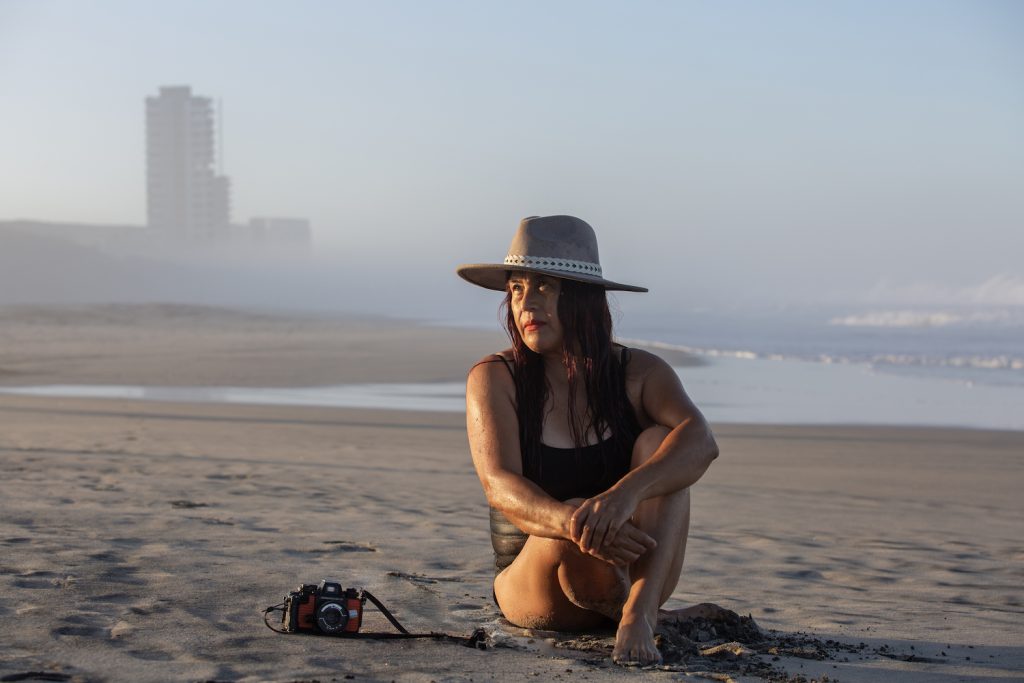Mexican artist Angélica Escoto sitting on the beach besides her Nikonos film camera with Tijuana in the background
