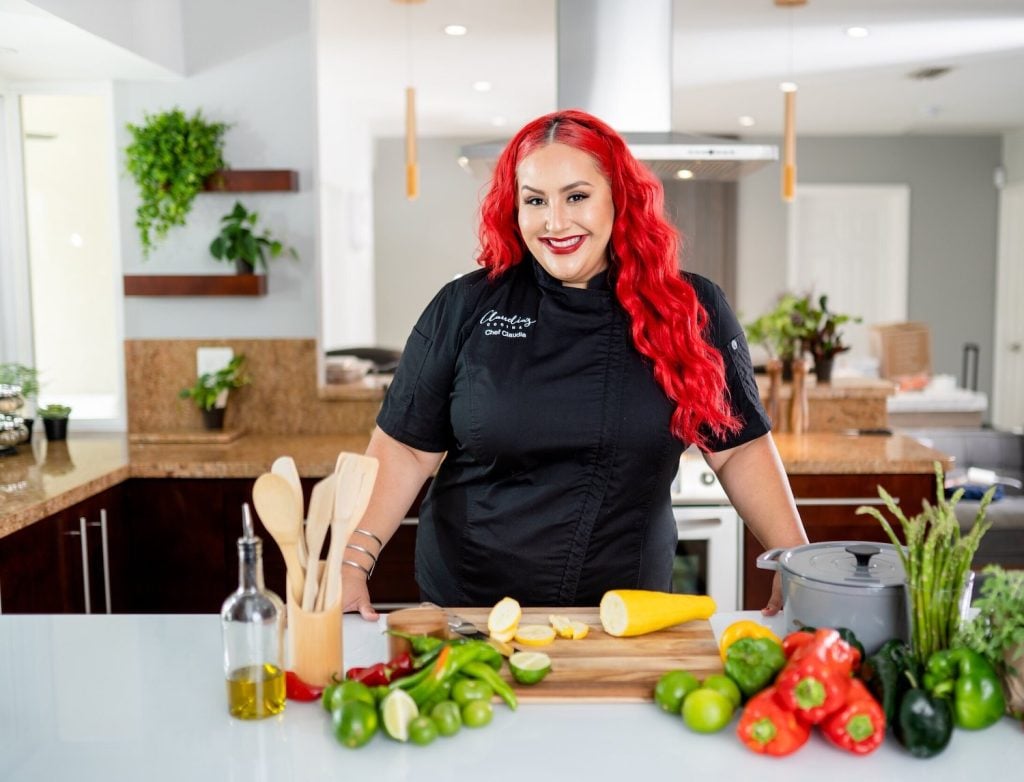 Celebrity chef Claudia Sandoval stands in a kitchen surrounded by fresh produce and kitchen utensils. 