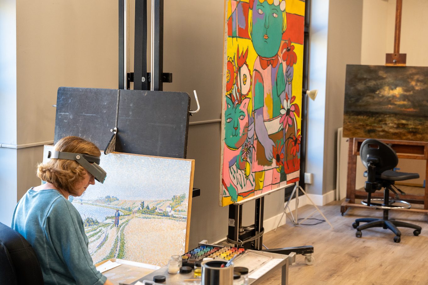 An art conservator works at an easel with a paint brush, paints,  and several other paintings in the background