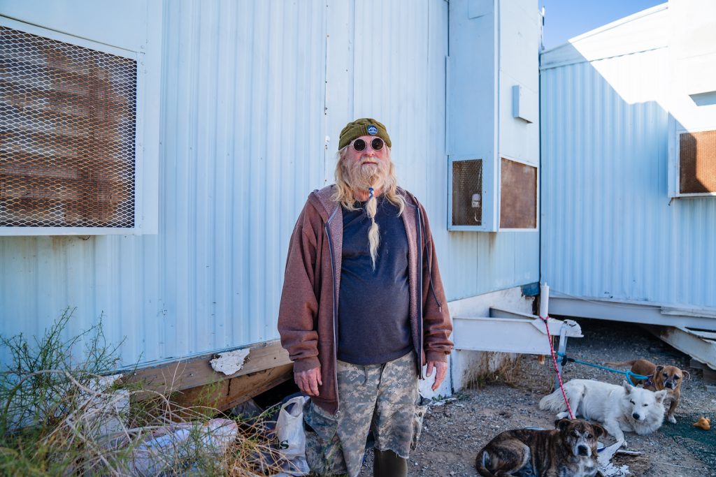 Veteran Tom Allen standing in front of his home in Slab City with his three dogs by his side