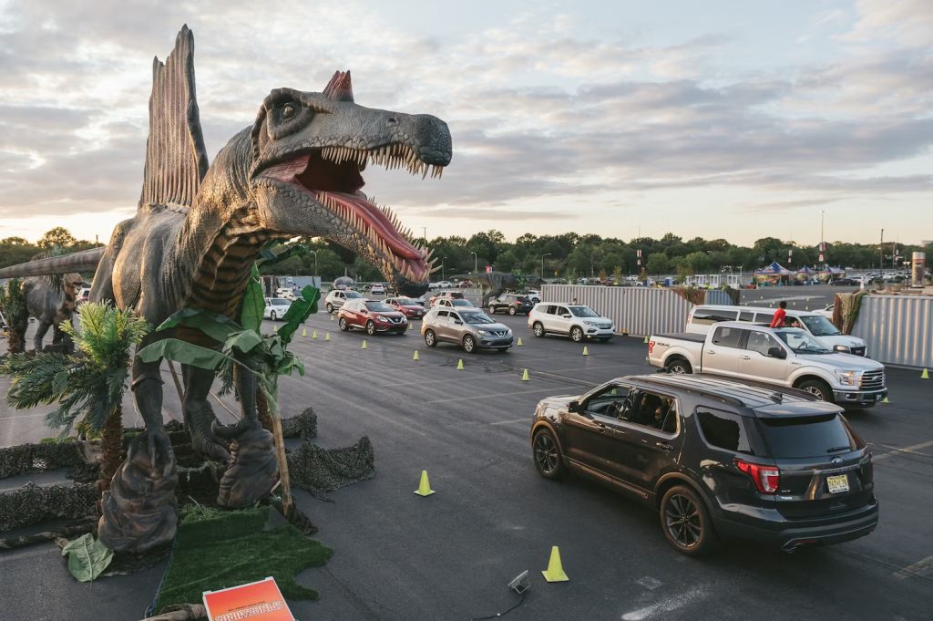 Jurassic Quest at the Del Mar Fairgrounds featuring a mid-sized car in a parking lot driving past a life-size replica of a dinosaur