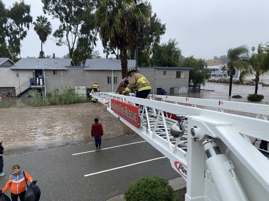 2024 San Diego flood relief fundraisers and resources for community support featuring a firefighter helping local families escape the flash flooding