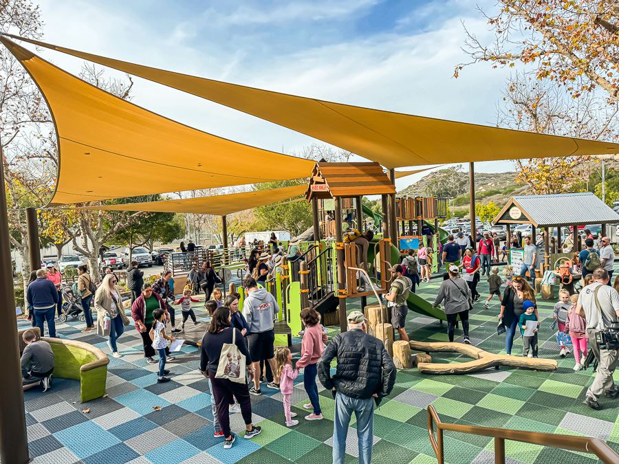Best new San Diego playgrounds and parks including Lake Poway Playground featuring a gathering of families and kids playing on the jungle gym underneath a shade covering