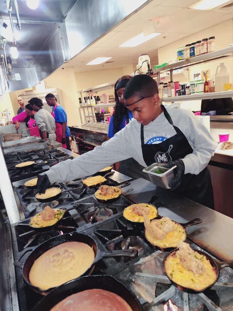 Mercedes Tiggs assists a young foster care student cook over a stove at a Eight16 Culinary Therapy session at the Joe and Vi Jacobs Center’s community kitchen