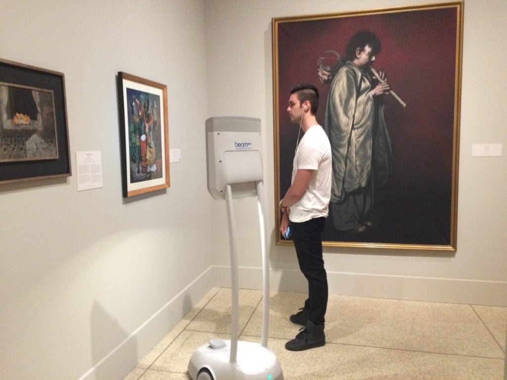 A man admiring art with earphones in and a Beam accessible screen nearby helping an impaired visitor