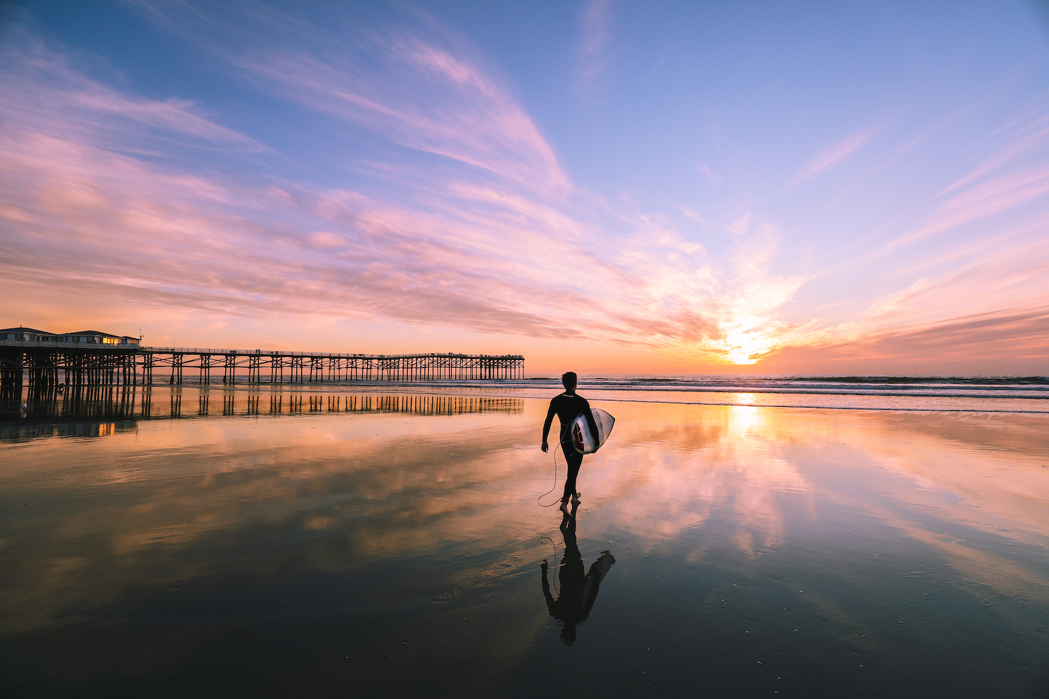 The best surf spots in San Diego featuring Pacific Beach's Crystal Pier with a surfer walking on the beach at sunset