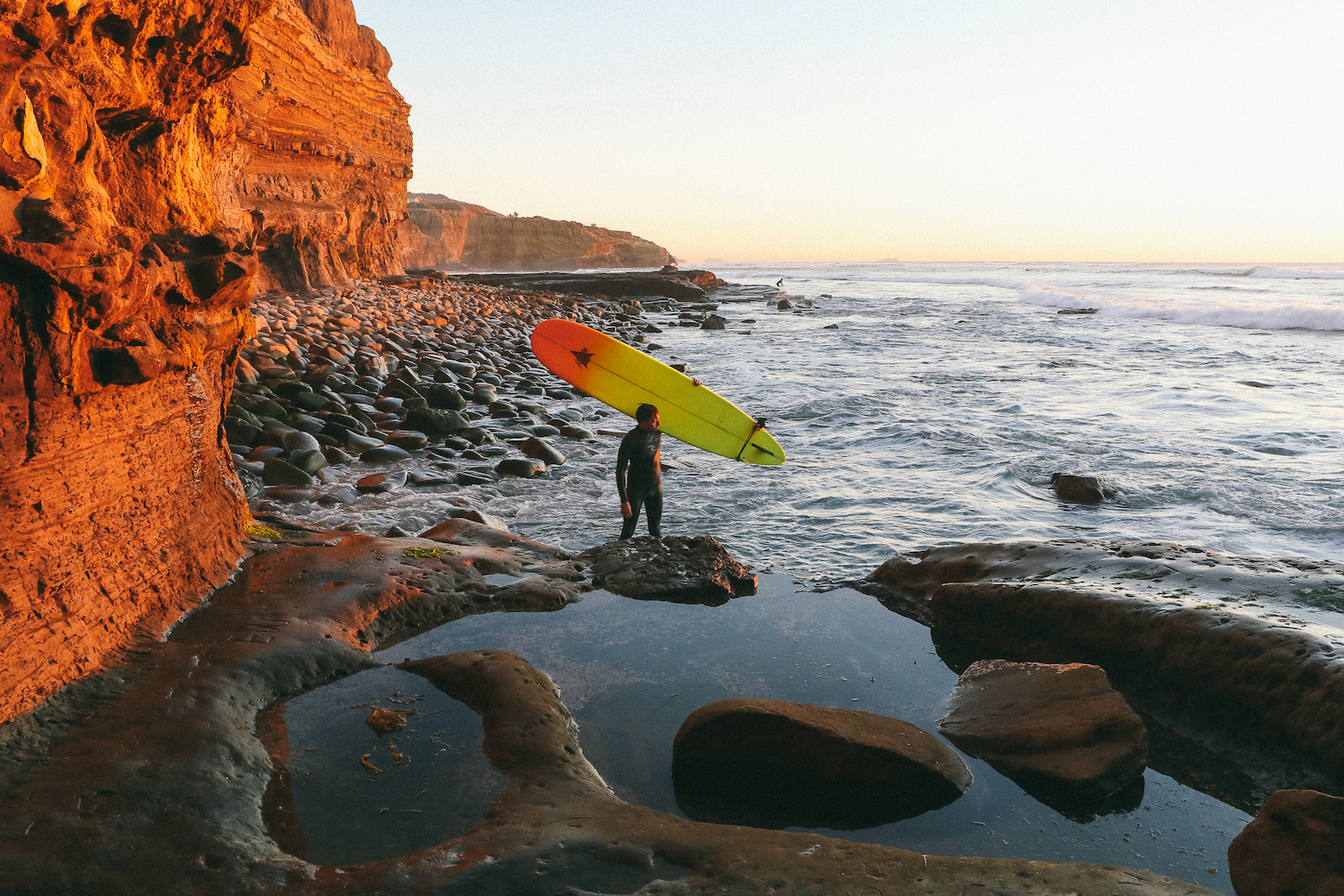 The best surf spots in San Diego featuring Sunset Cliffs, Point Loma with a surfer carrying his longboard surfboard at sunset 